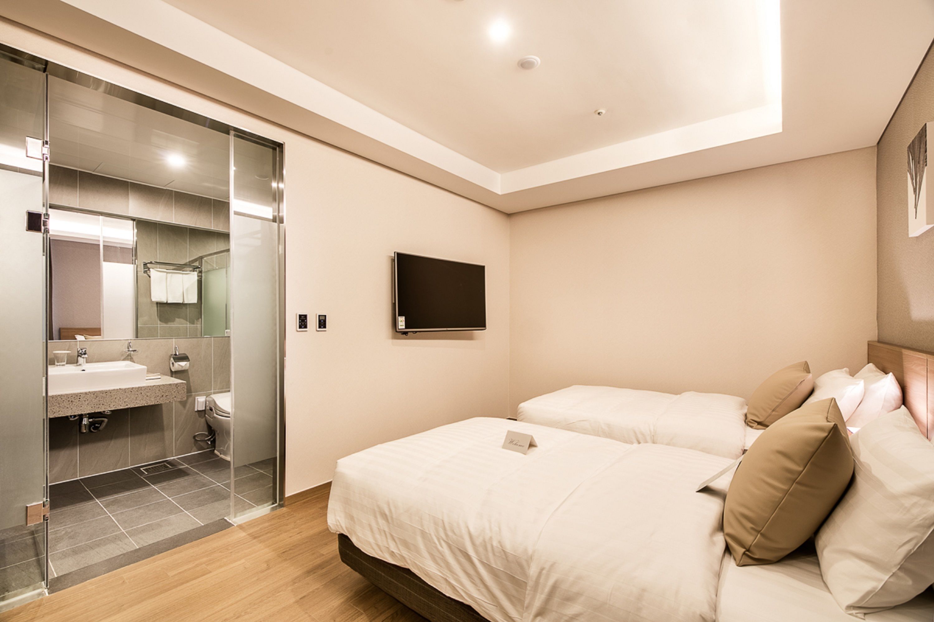 A modern hotel room with a glass shower enclosure, a flat-screen TV mounted on the wall, and a bed with beige bedding.