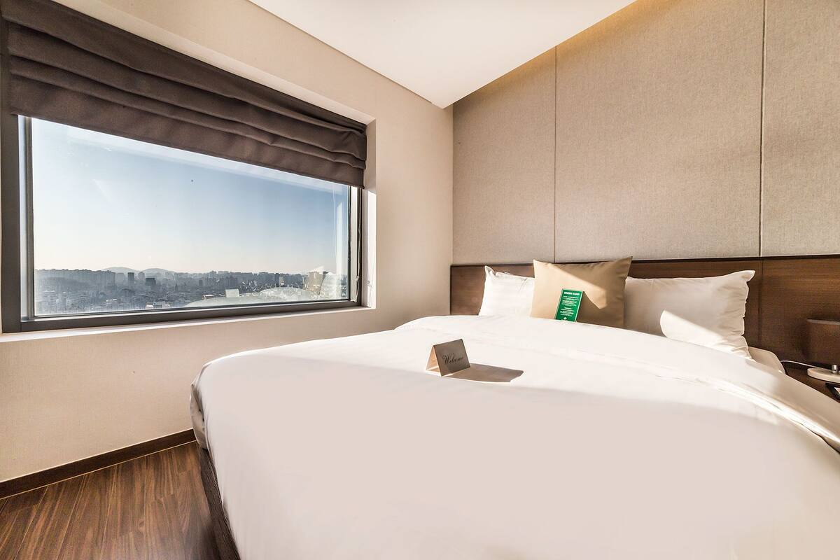 A hotel room with a large bed, a city view through a window, and a bedside table with a lamp.