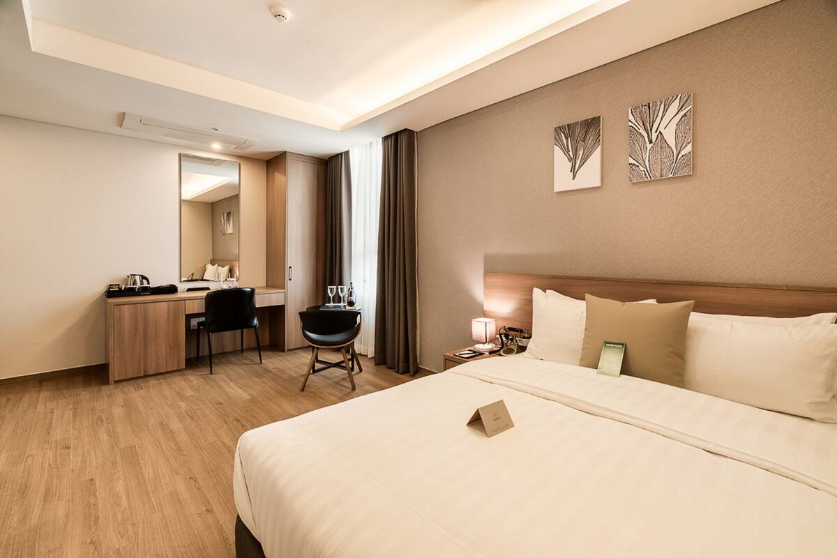 A hotel room with a large bed, a desk, a chair, and a nightstand with a lamp.