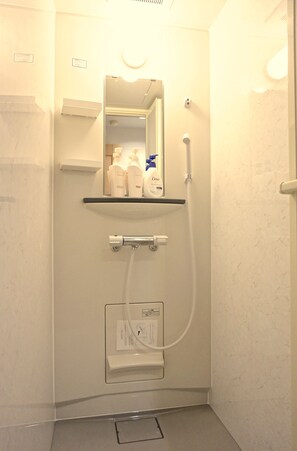 A small, well-lit bathroom with a mirror, a shelf with toiletries, a handheld showerhead, and a wall-mounted faucet. - Hostel Rakutsuki (Osaka)