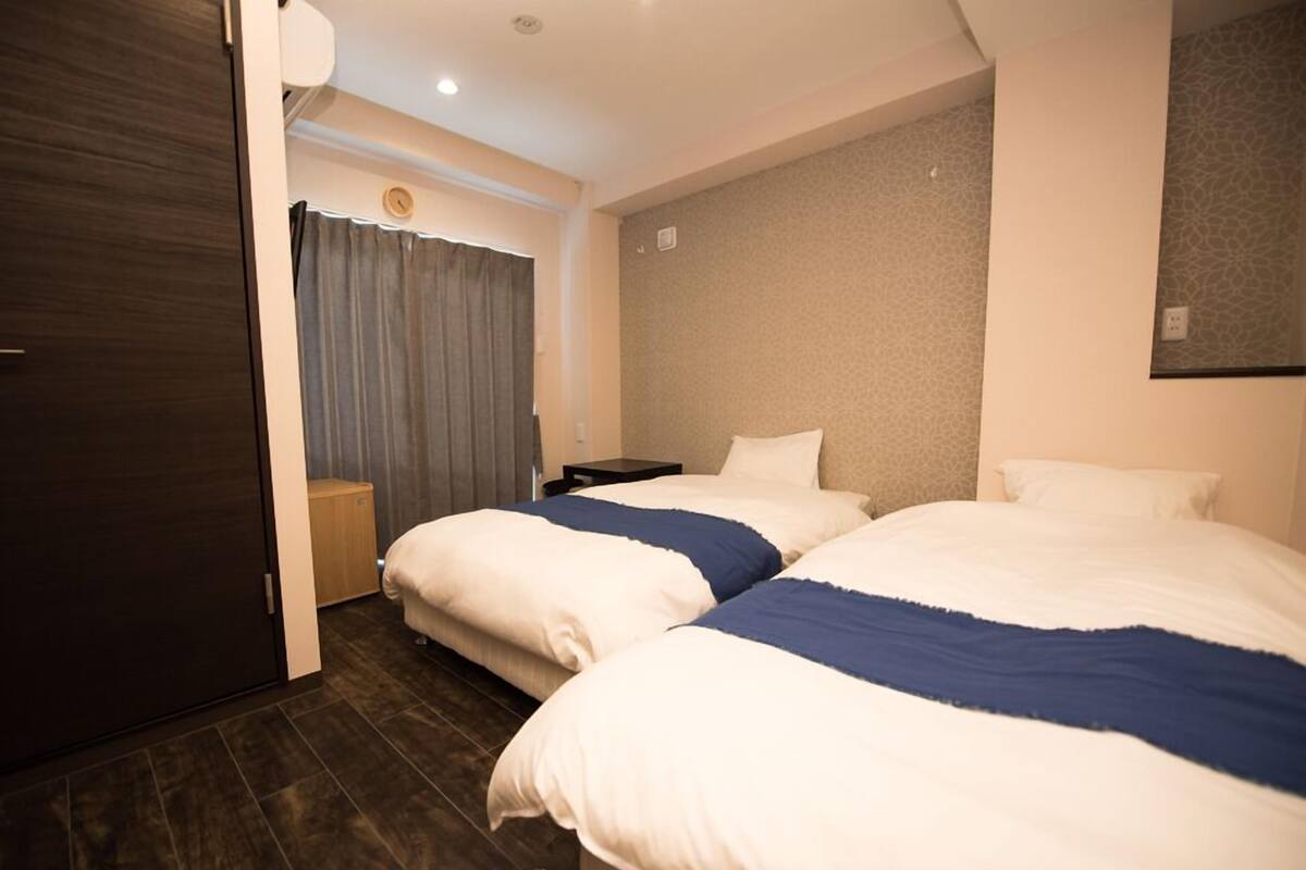 Una habitación de hotel con dos camas, un armario de madera, una mesita de noche pequeña y una cortina.