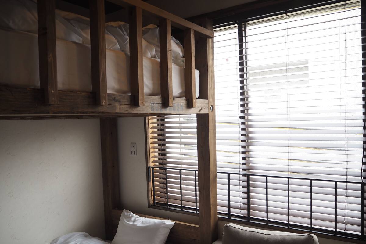 A bunk bed with a wooden headboard, a single bed with a pillow, and a window with blinds.