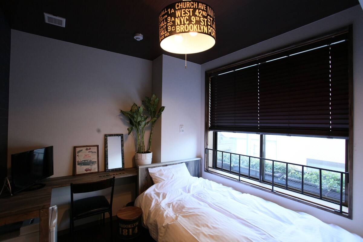 A bedroom with a bed, a desk, a chair, a television, a potted plant, and a window with blinds.