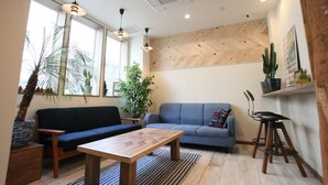 A living room with a wooden coffee table, a blue sofa, a blue armchair, and a small wooden stool. There are two pendant lights, a large window with a view of palm trees, and a wooden herringbone wall panel. - North Front Hostel (Fukuchiyama)