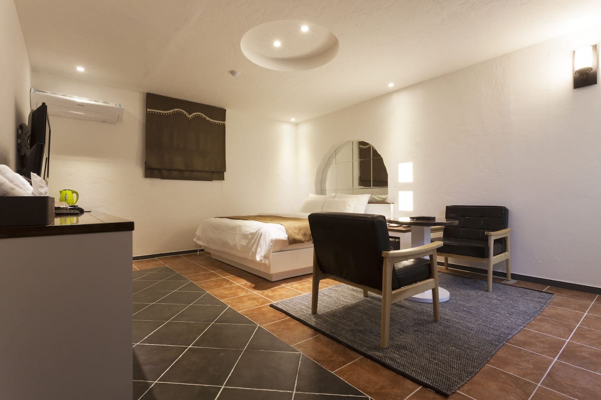 A hotel room with a bed, a desk, a chair, a mirror, and an air conditioning unit.