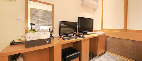 A hotel room with a wooden desk, two monitors, a keyboard, and a telephone.