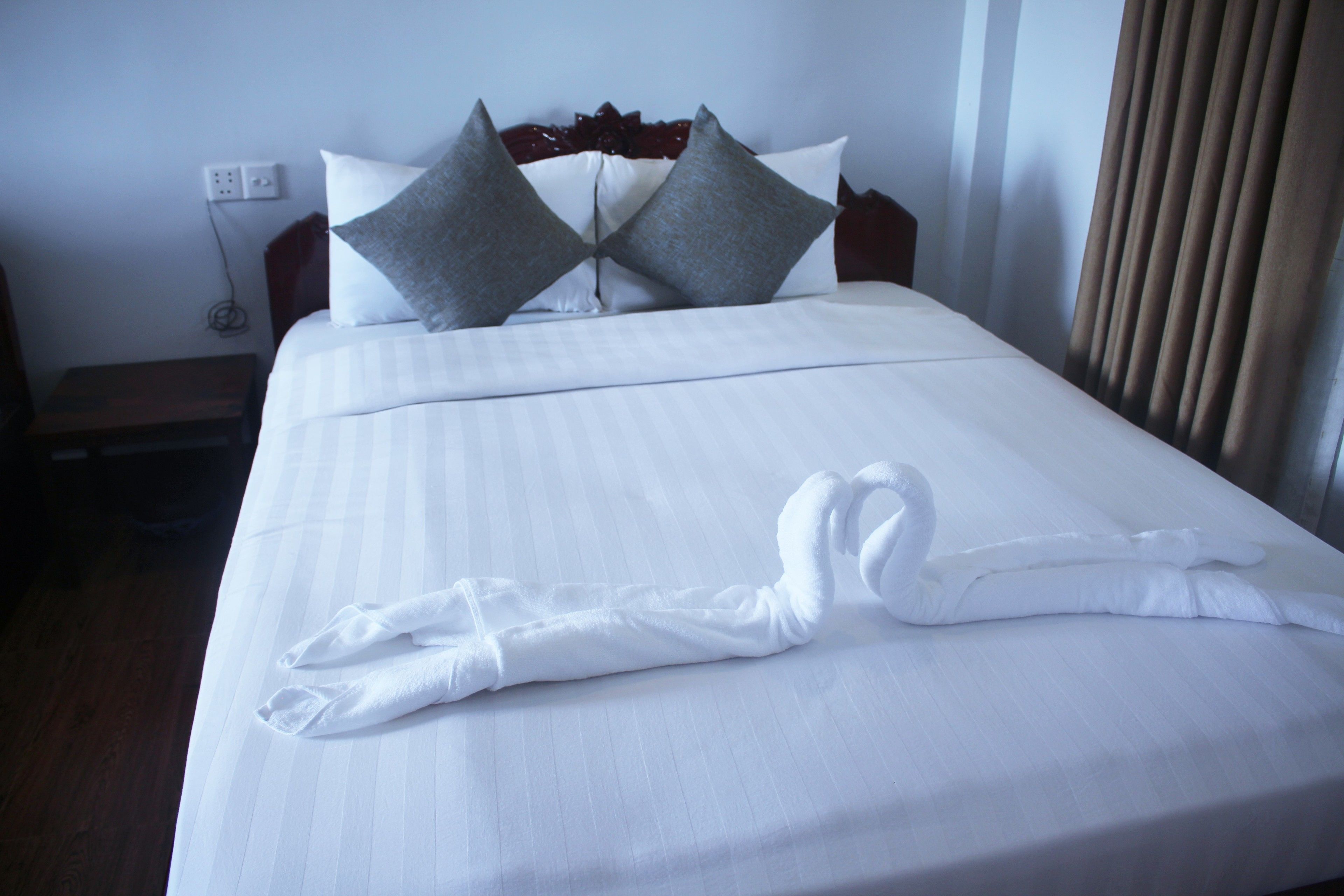 A neatly made bed with a towel and a swan-shaped towel arrangement, a wooden headboard, and a power outlet on the wall.