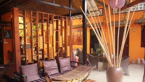 A wooden bench with cushions, a large vase with bamboo sticks, and a wicker chair in a room with orange walls and a tiled floor. - Ramchang Guesthouse (Battambang)