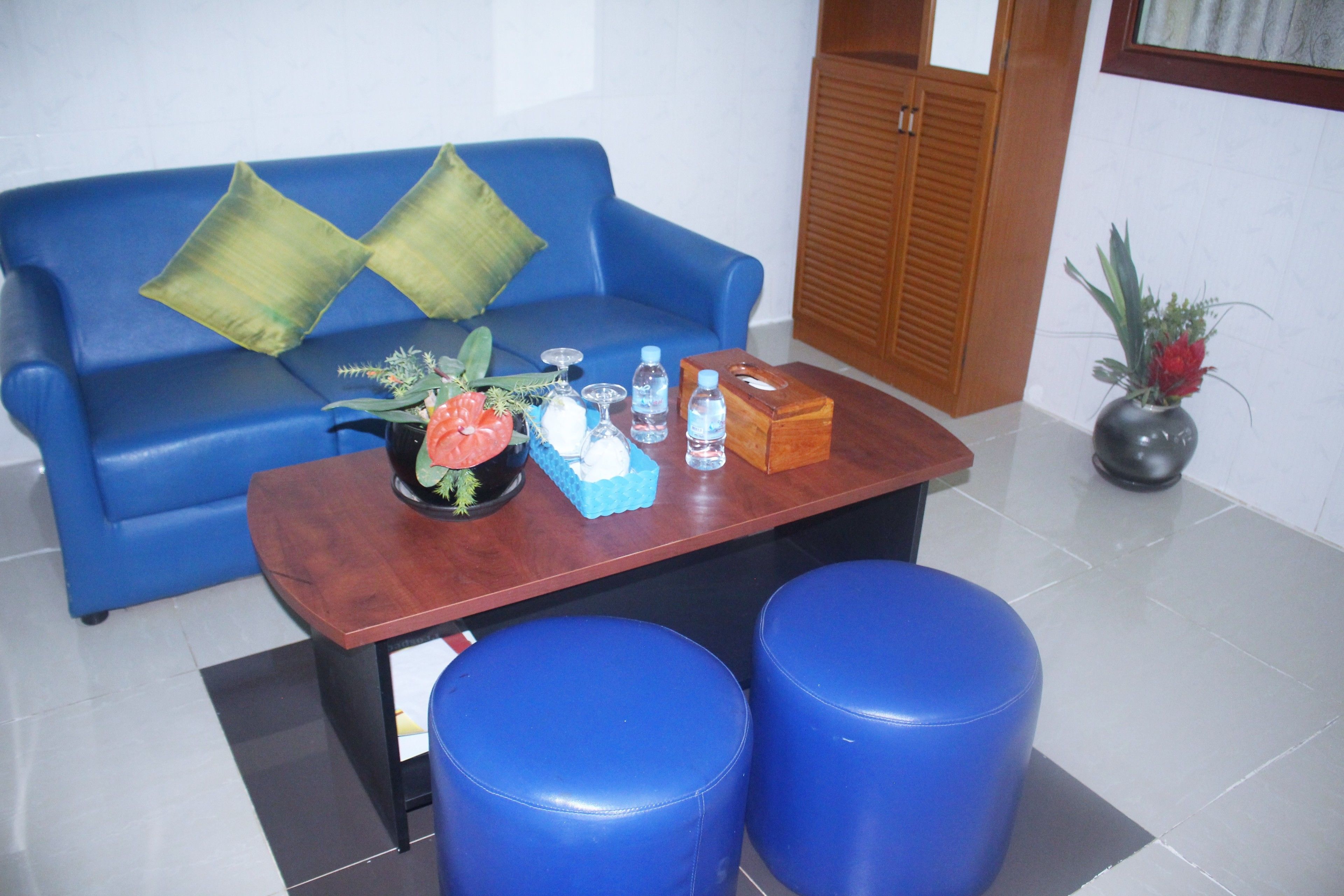 Un sofá azul con cojines verdes, una mesa de centro de madera con un jarrón de flores, dos taburetes azules y una planta en maceta.