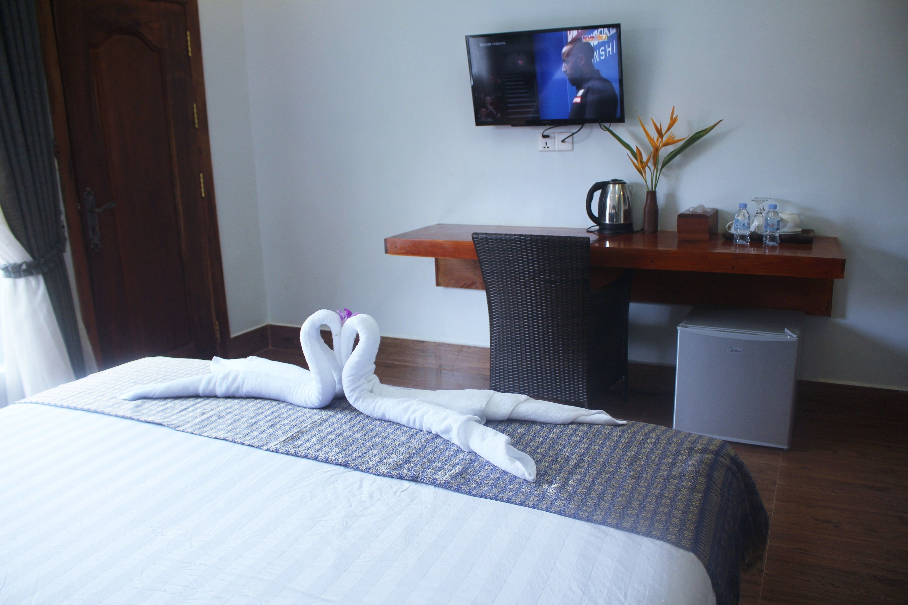 Ein Hotelzimmer mit einem Bett, einem an der Wand befestigten Fernseher, einem Schreibtisch mit Stuhl, einer kleinen Kühlbox und einer Vase mit Blumen.