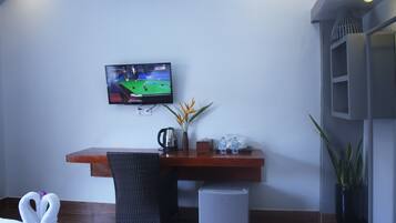A room with a television displaying a pool game, a wooden desk, a chair, a small refrigerator, and two potted plants.