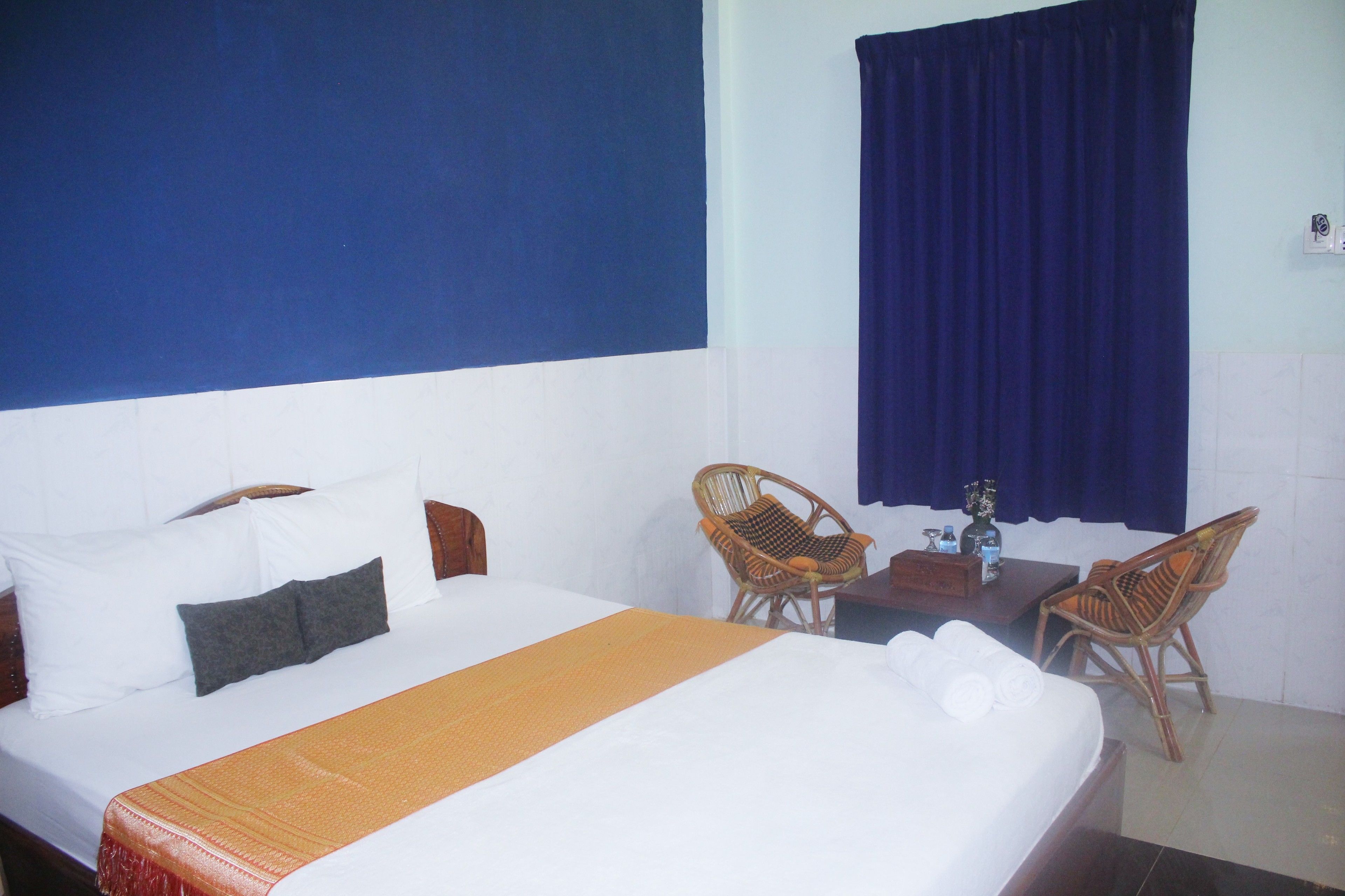 A hotel room with a bed, two wicker chairs, a small table, and a blue curtain.