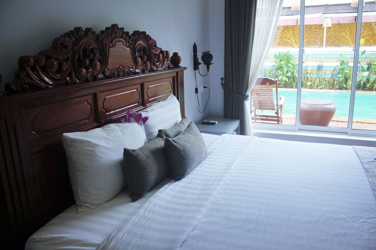 A hotel room with a large bed, a wooden headboard, and a view of a swimming pool through a window.