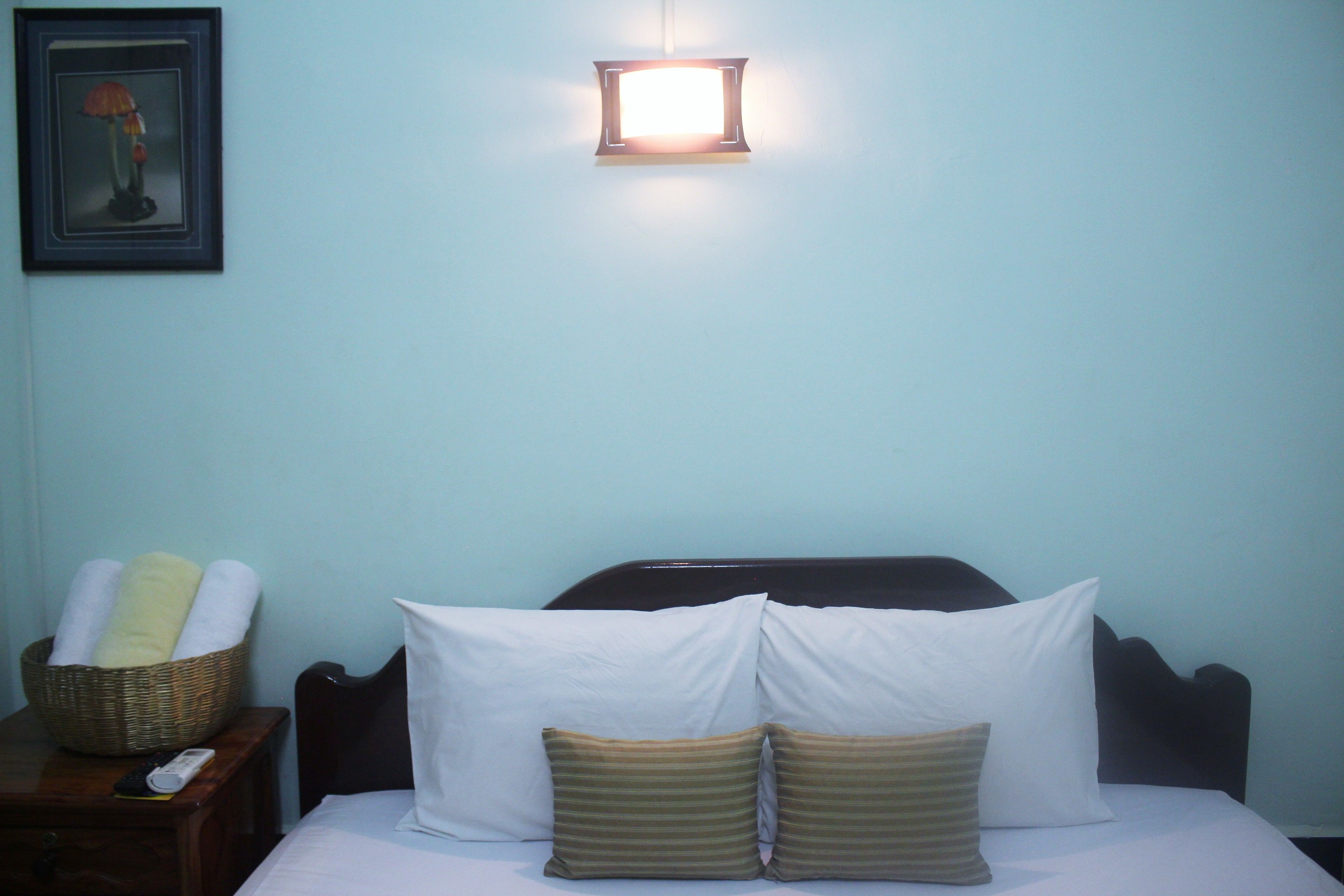 A neatly made bed with white and striped pillows, a remote control, and a basket of towels on a wooden nightstand.