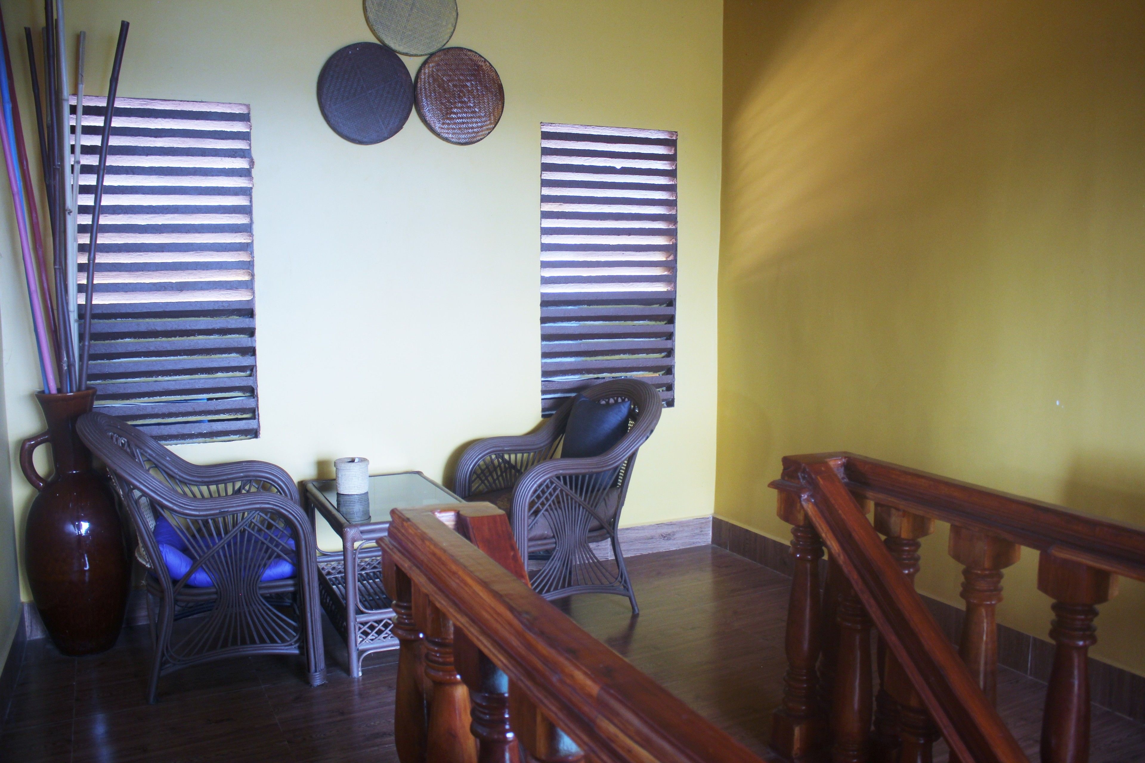Um quarto com uma mesa de madeira, duas cadeiras de vime, um vaso grande e um corrimão de madeira.