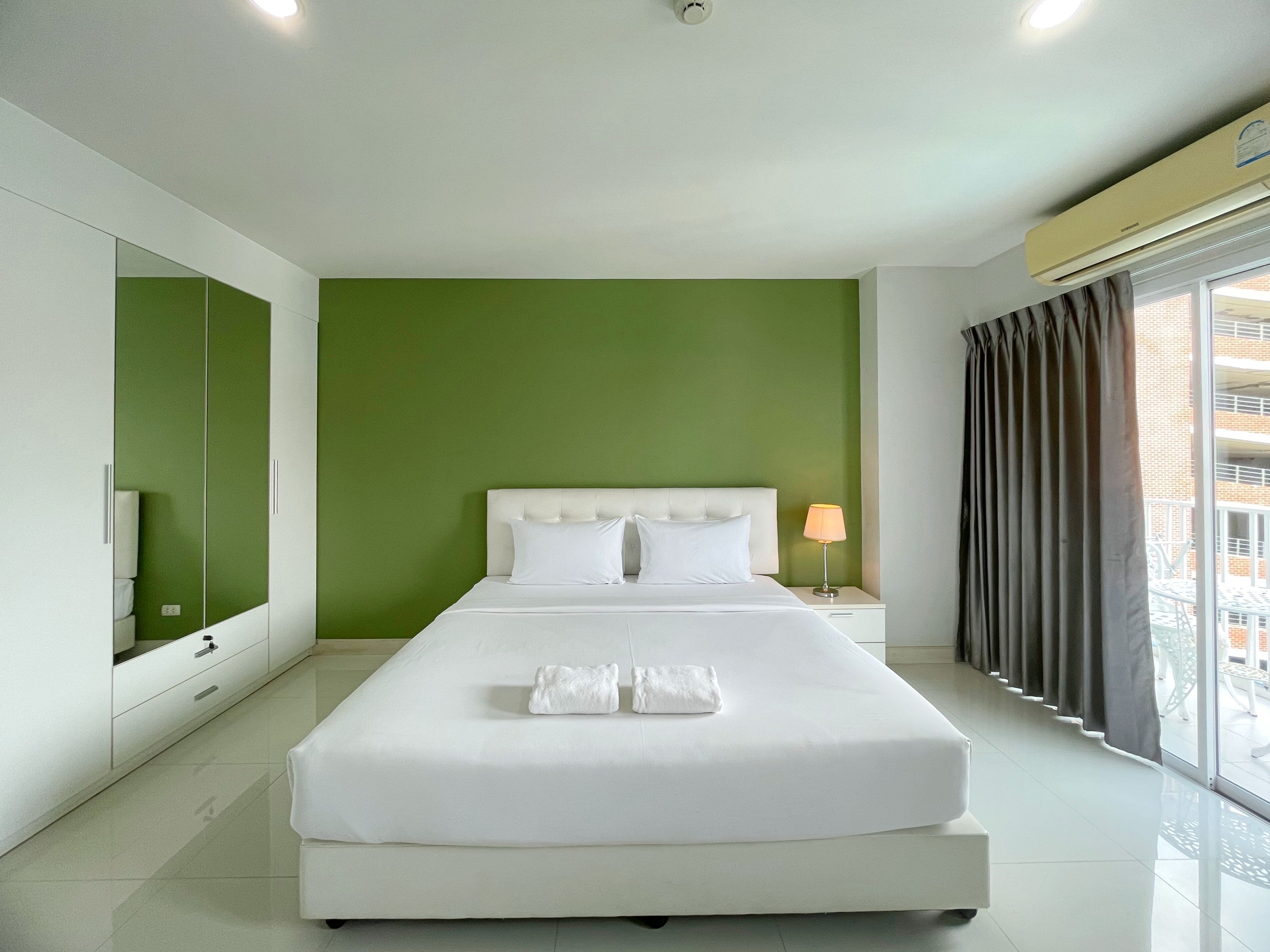 Una habitación de hotel moderna con una cama grande, paredes verdes, una ventana con cortinas y una mesita de noche con lámpara.