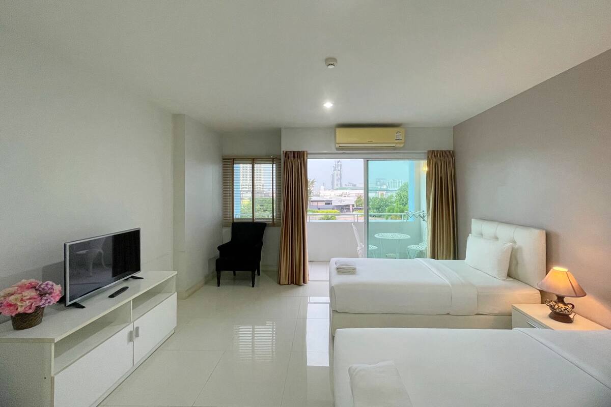A hotel room with a bed, a TV, a desk, and a chair. There is a balcony with a view of buildings.