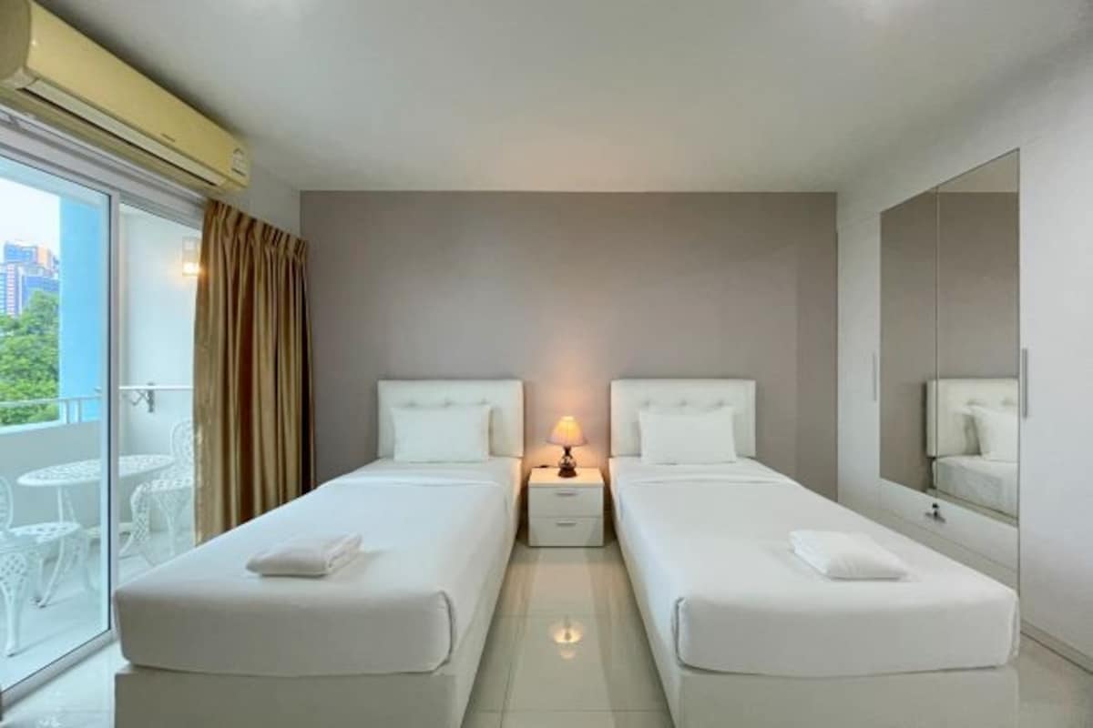 A hotel room with two beds, a small bedside table with a lamp, a large mirror, and a sliding glass door leading to a balcony with outdoor furniture.