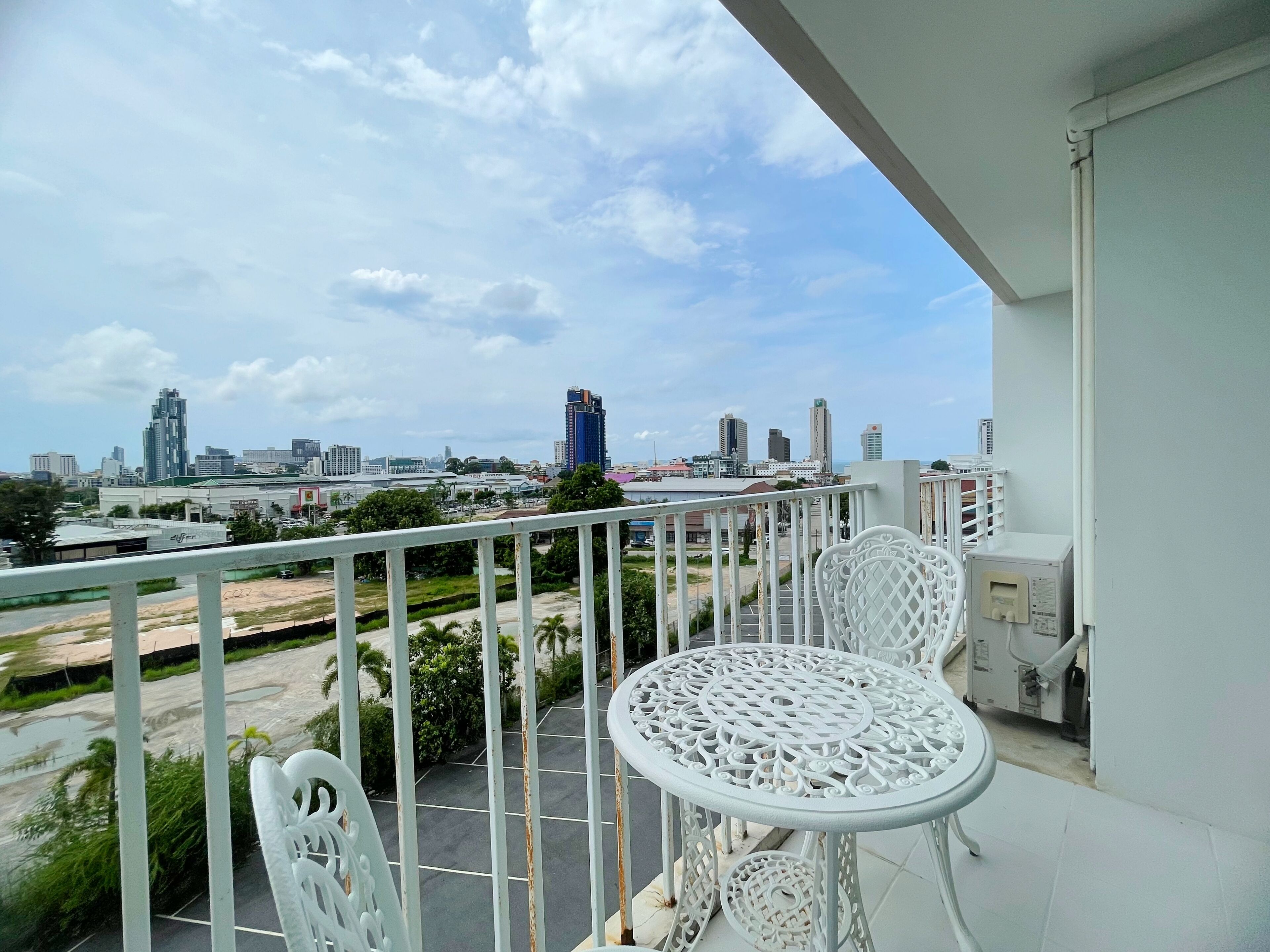 A balcony with white metal railings, a white metal table, and two white metal chairs. In the background, there is a cityscape with various buildings and a clear sky.