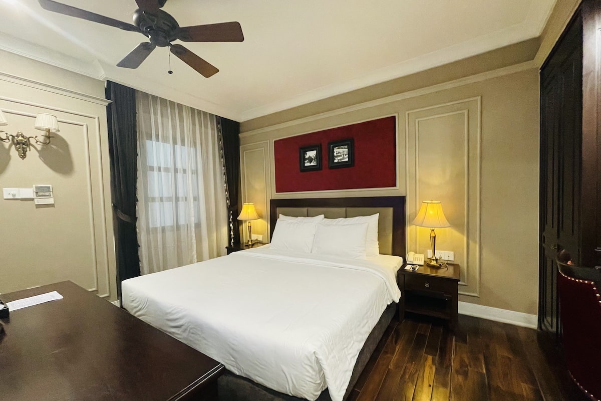 Habitación de hotel con una cama, dos mesitas de noche con lámparas, un ventilador de techo, un escritorio de madera y un cuadro con marco rojo en la pared.