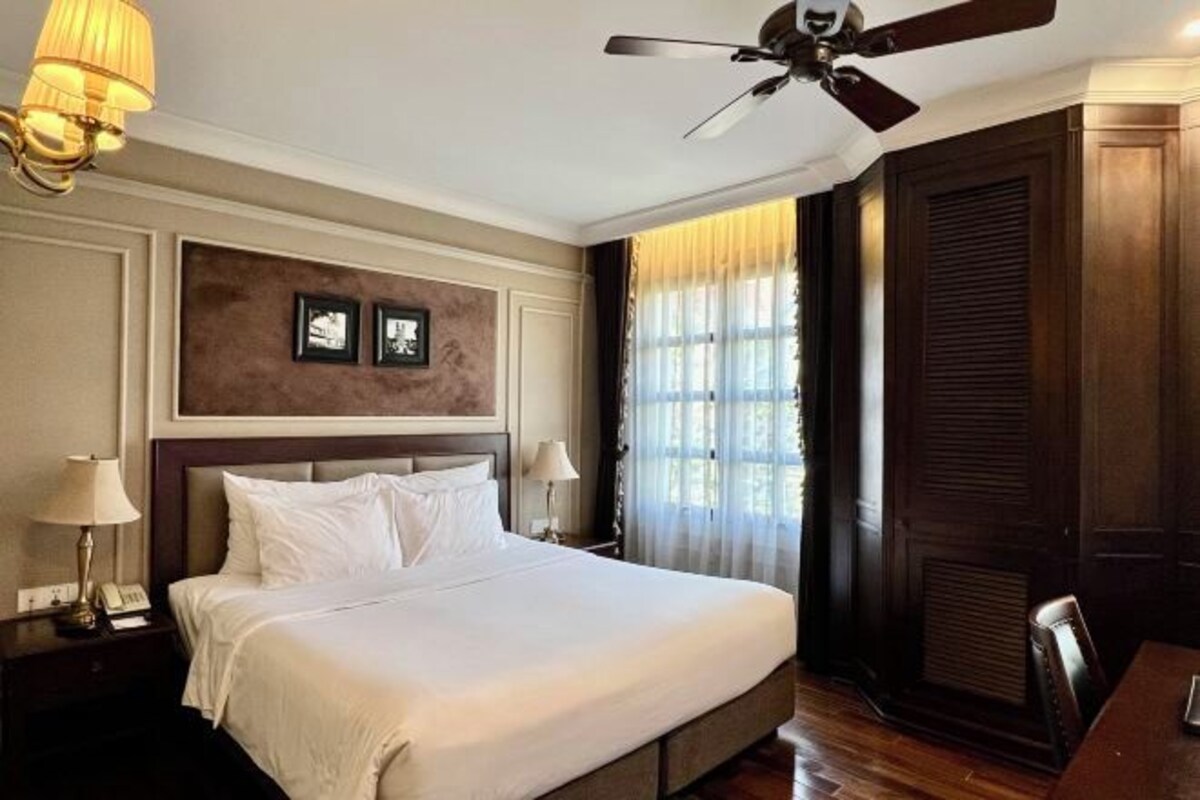 A hotel room with a large bed, a ceiling fan, a nightstand with a lamp, and a window with curtains.