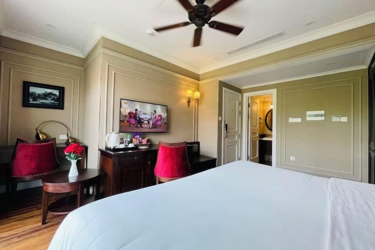 Une chambre d’hôtel avec un grand lit, un bureau avec une chaise, une télévision et un ventilateur de plafond.