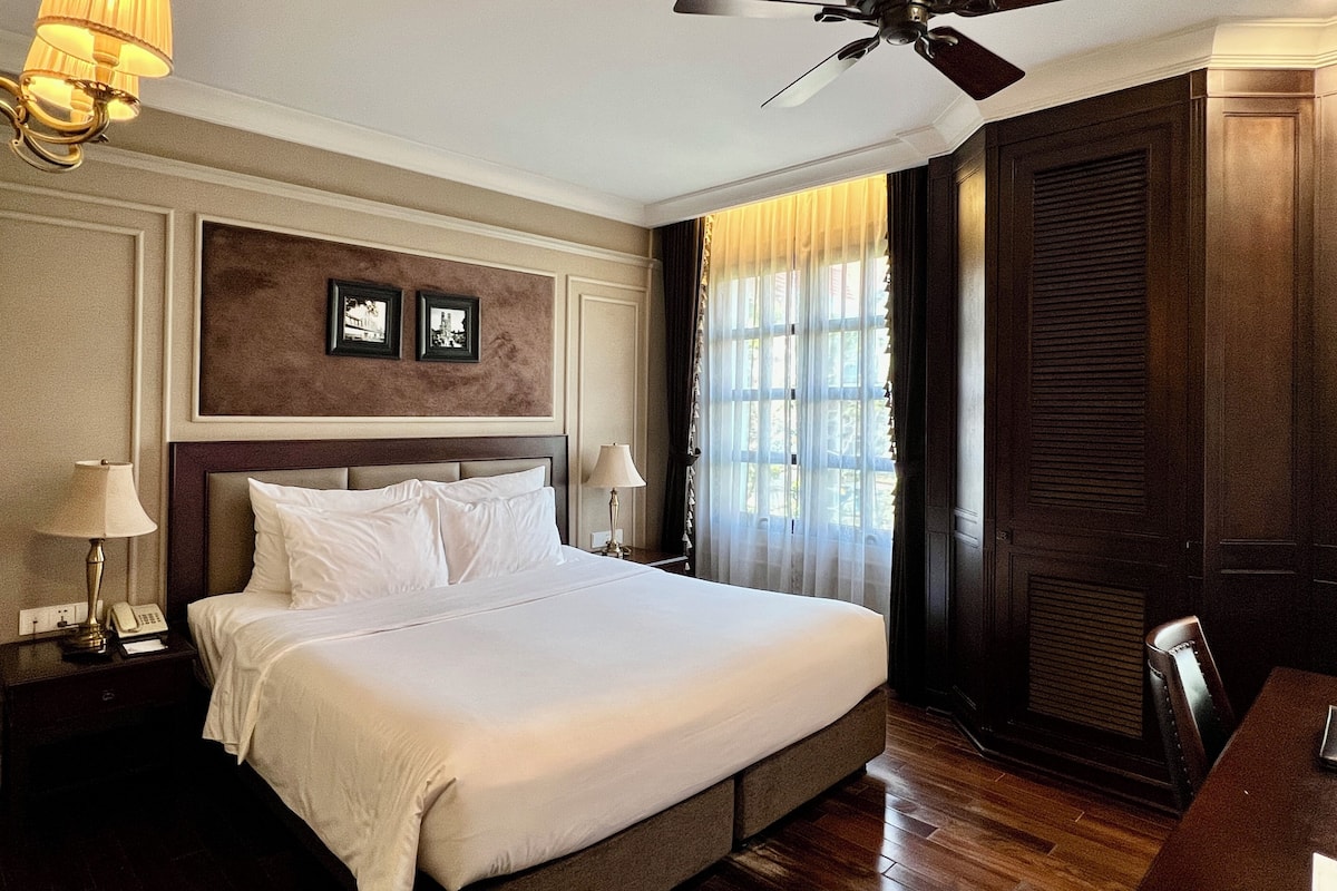 Habitación de hotel con una cama grande, ventilador de techo, mesita de noche con lámpara, escritorio con silla y ventana con cortinas.