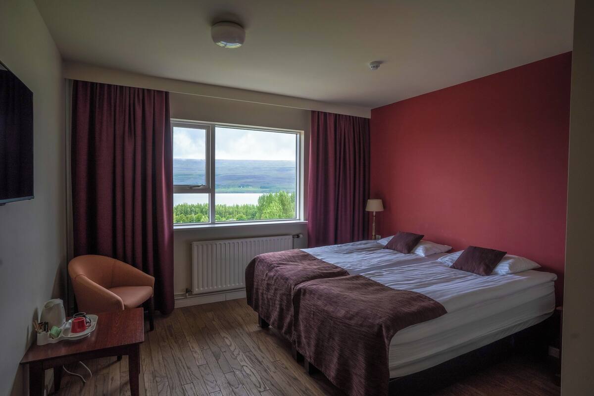 Ein Hotelzimmer mit Bett, Sessel, Tisch mit Gegenständen, Fenster mit Vorhängen und Blick auf die Landschaft.