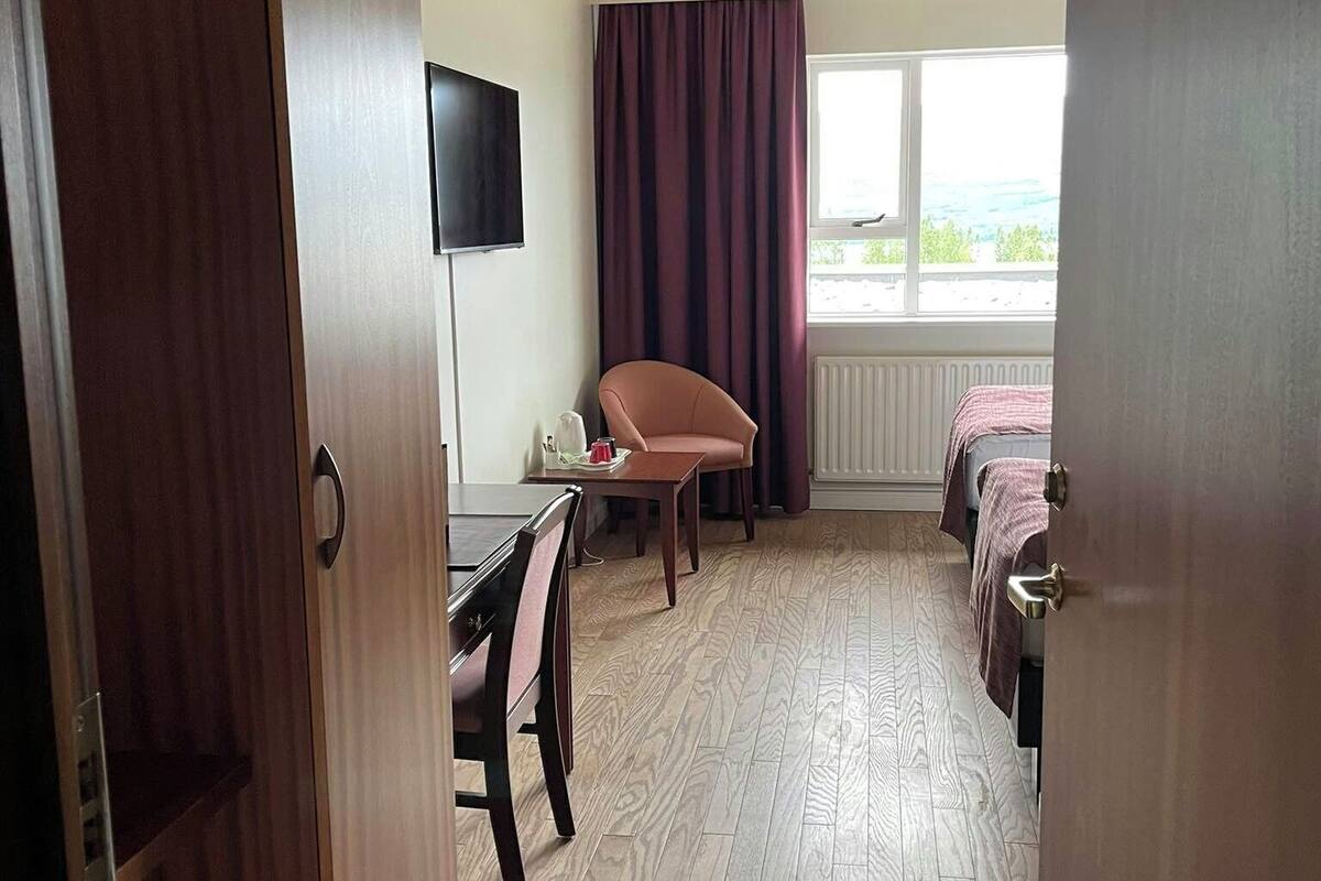 A hotel room with a wooden floor, a desk with a chair, a bed, a TV mounted on the wall, and a window with curtains.