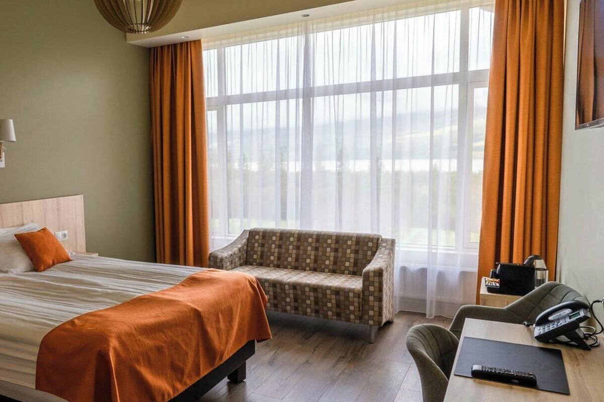 Ein Hotelzimmer mit einem Bett, einer Couch, einem Schreibtisch mit Telefon und Fernbedienung sowie einem großen Fenster mit Vorhängen.