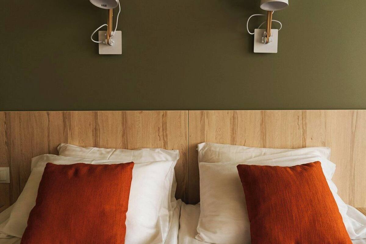Ein Hotelzimmer mit zwei Betten, jeweils mit weißer Bettwäsche und einem einzelnen orangefarbenen Kissen. Zwei Wandlampen mit weißen Schirmchen sind angebracht.