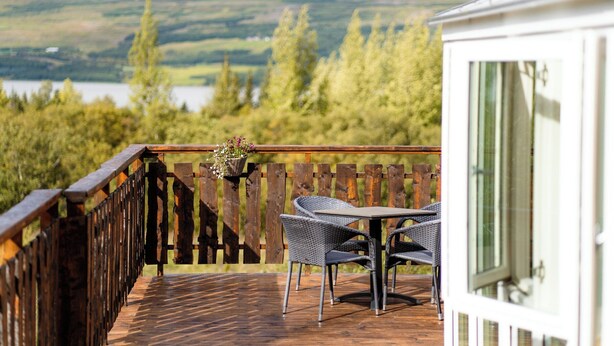 Um deck de madeira com mesa e cadeiras, com vista para um lago e colinas arborizadas.