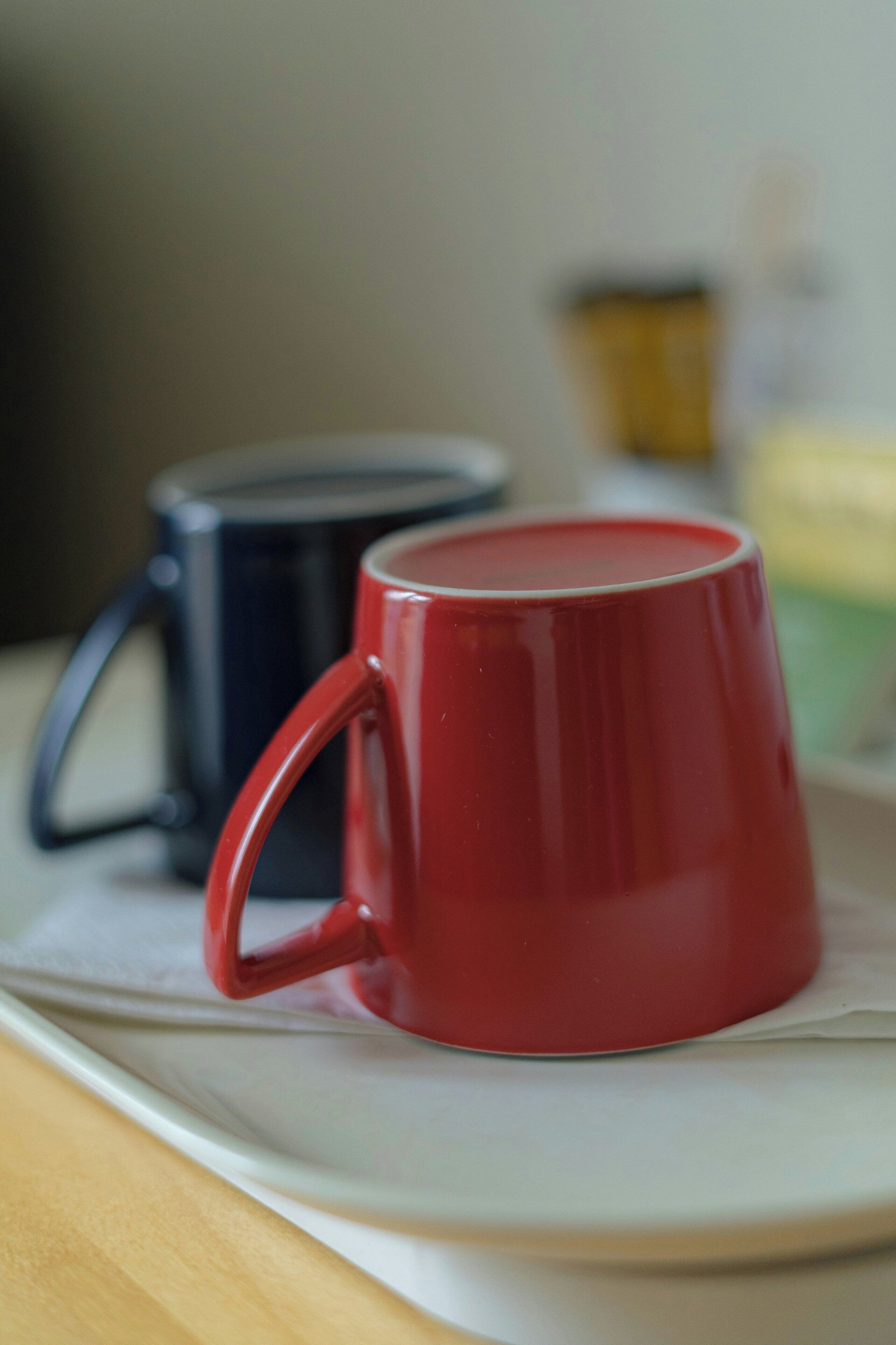 Una taza roja sobre un plato blanco, con otra taza azul al fondo.