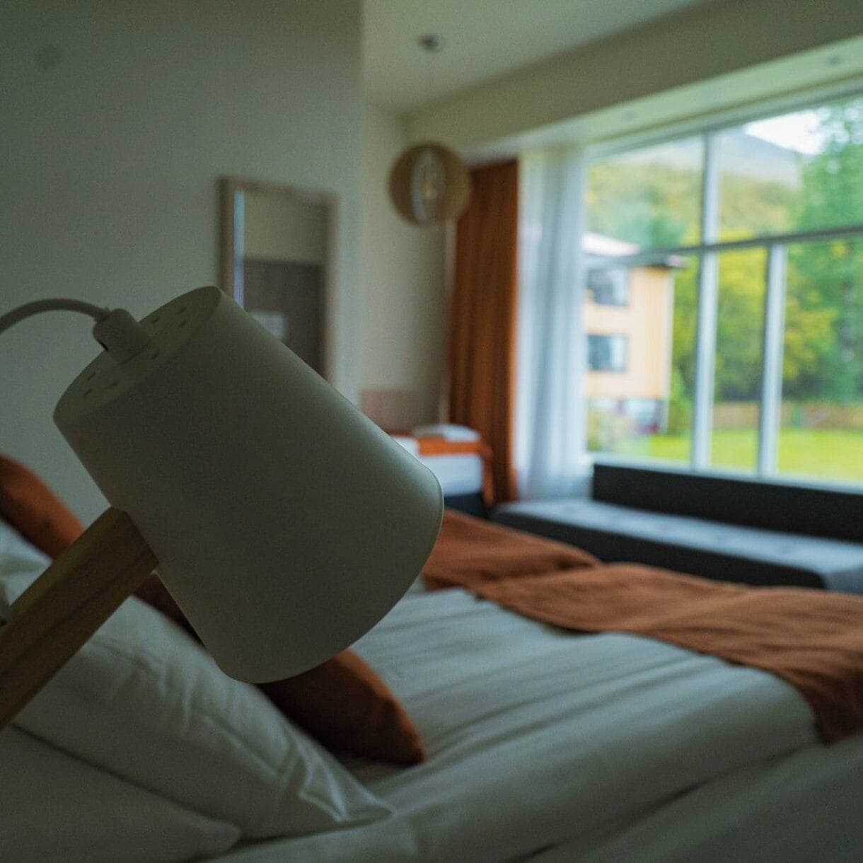 A bed with white bedding and a brown blanket, a bedside lamp, a window with a view of greenery, and a clock on the wall.