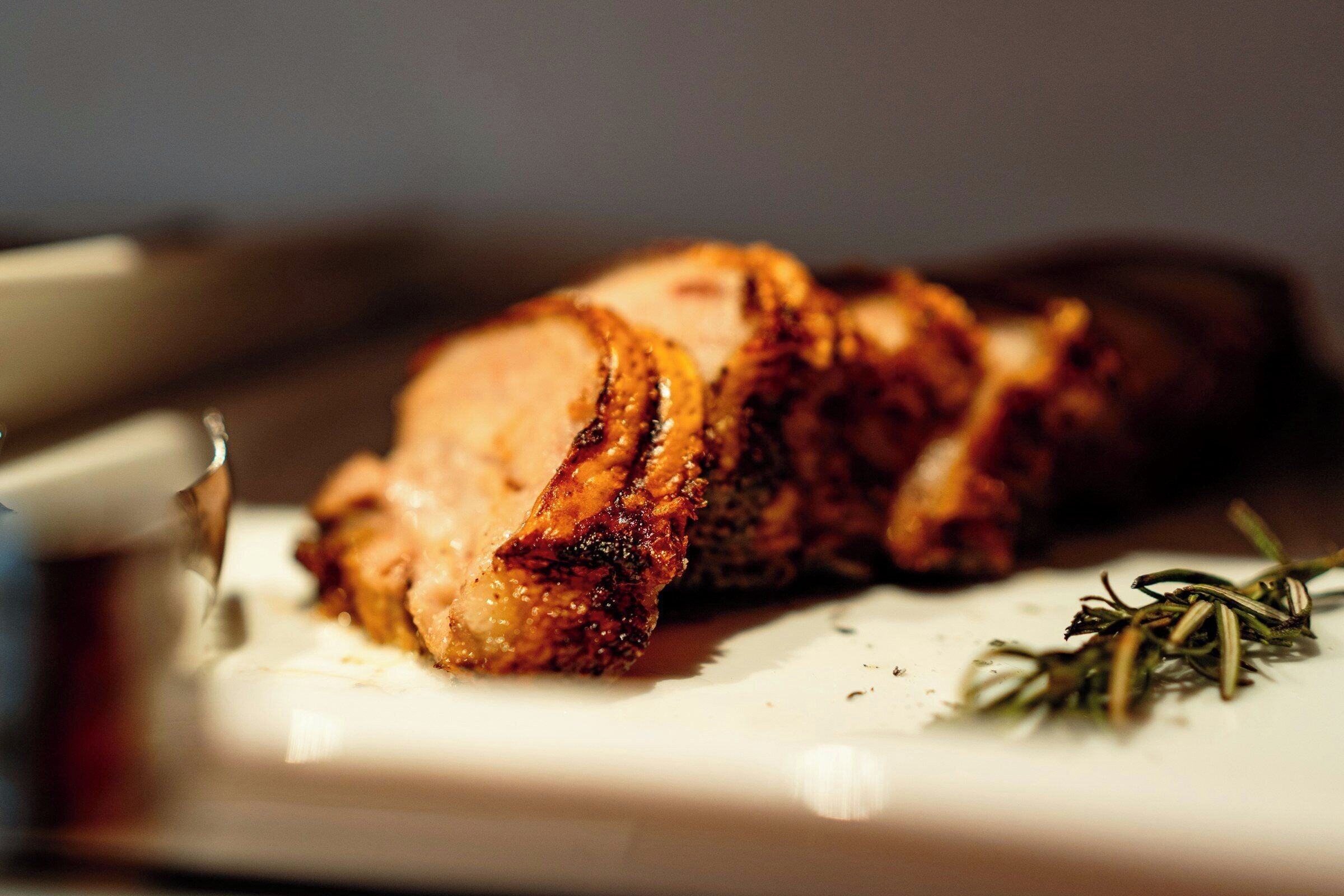 A close-up of sliced meat with seasoning, a knife, and a sprig of rosemary on a white plate.