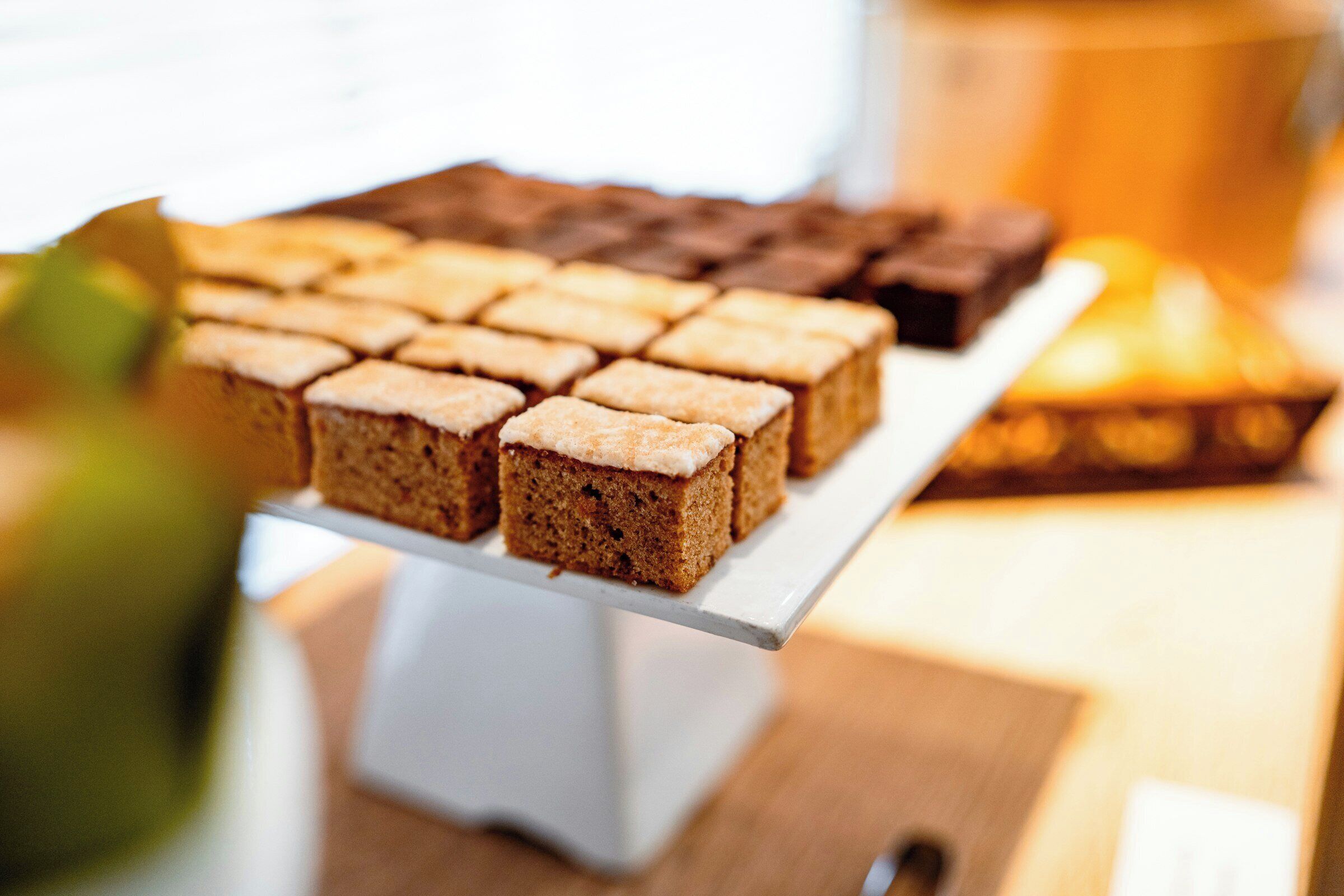 En hvid kagefad med flere firkantede desserter, sandsynligvis brownies, på et træbord.