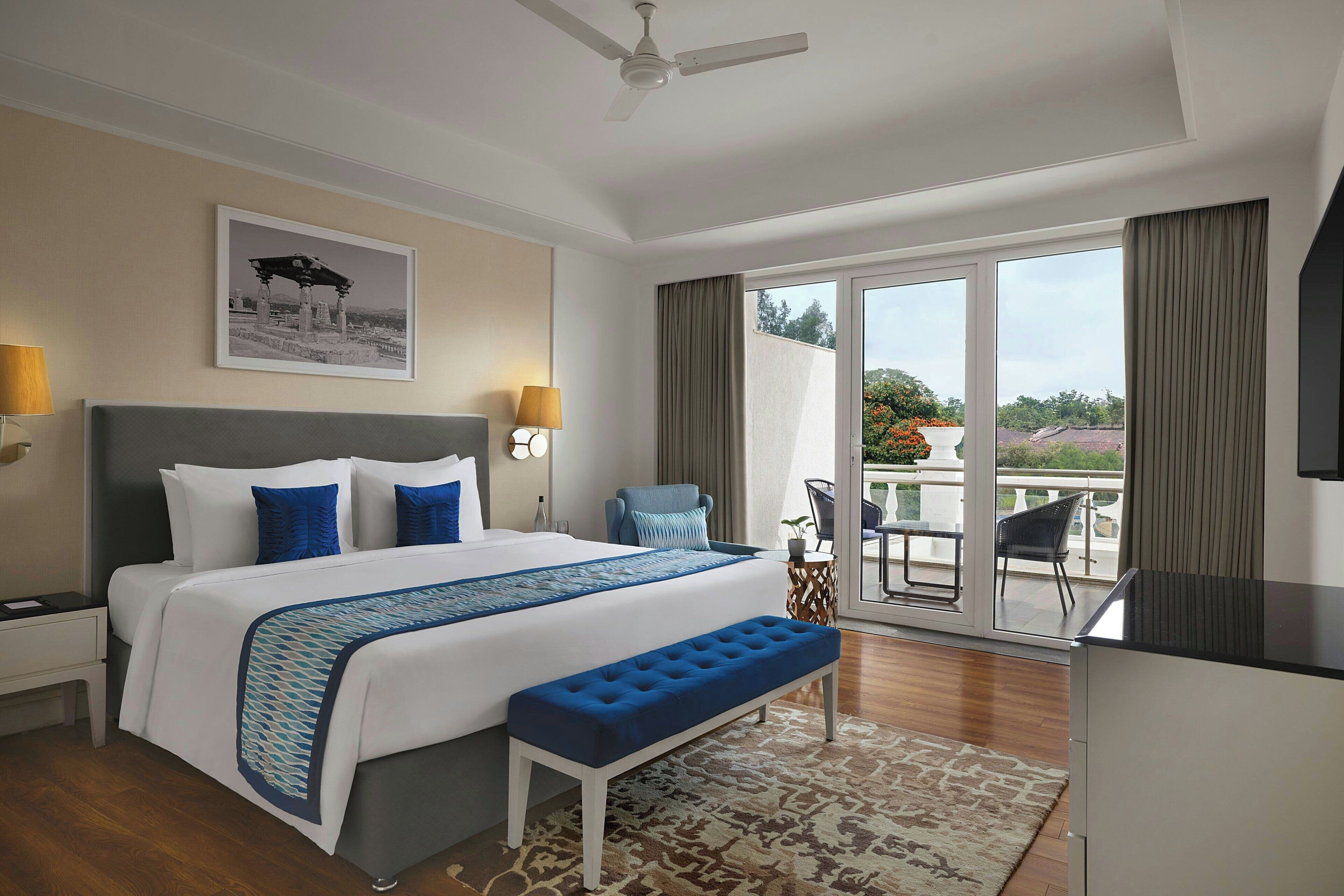 A hotel room with a large bed, a bench, a desk, a chair, and a balcony with a view.