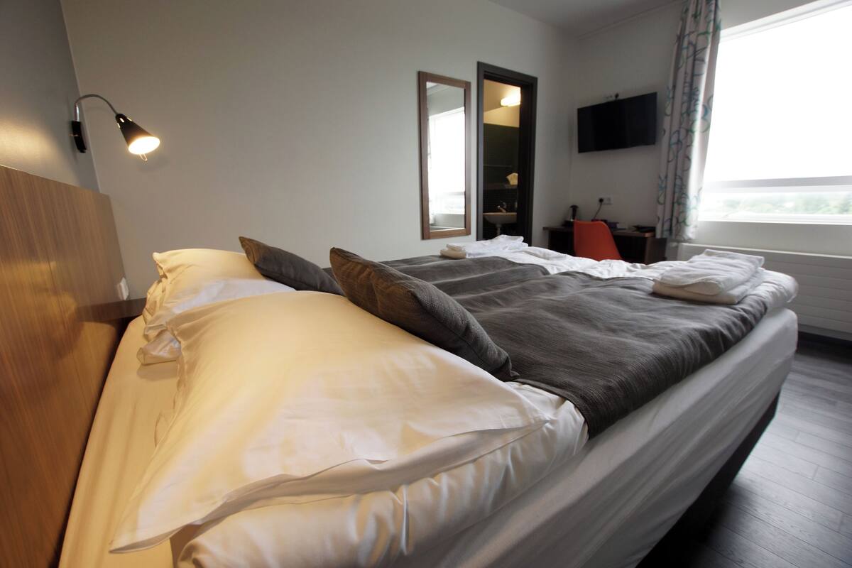 Ein Hotelzimmer mit Bett, Nachttischlampe, Spiegel, Fernseher und Fenster.
