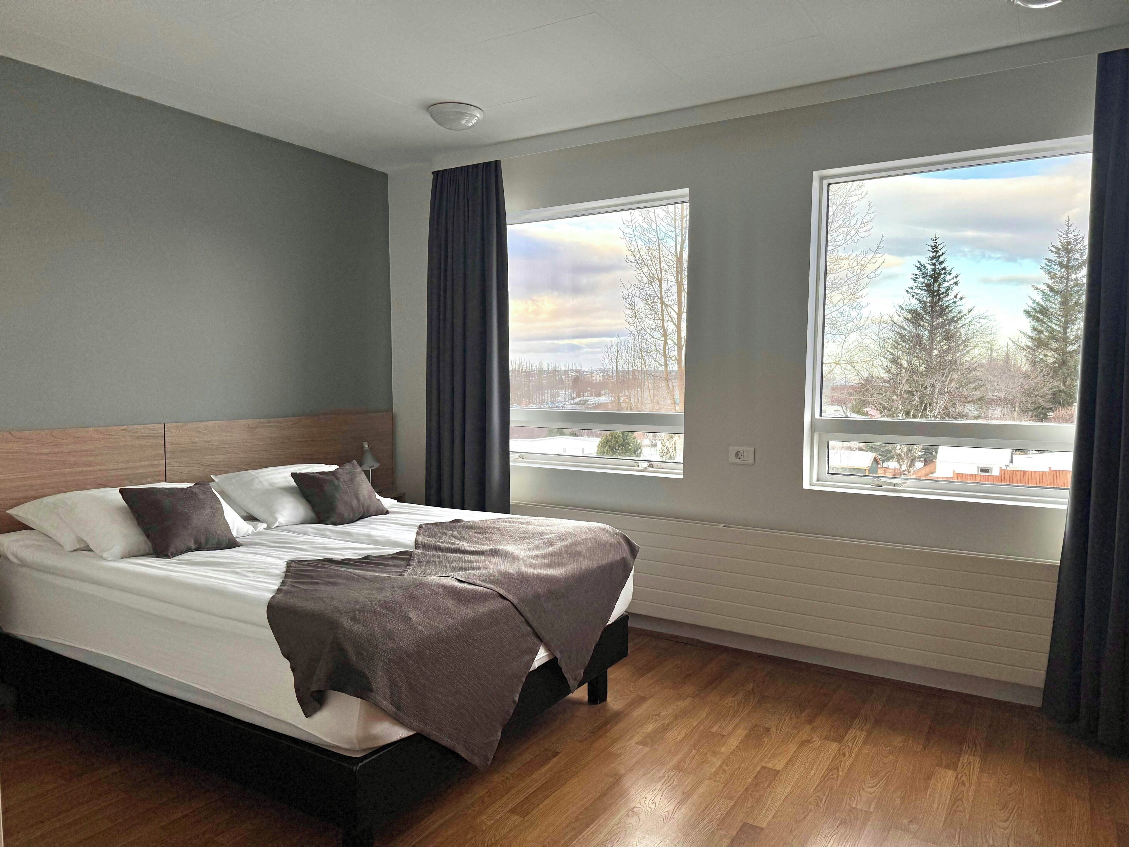 A modern hotel room with a large bed, two windows with curtains, a ceiling light, and a view of trees and buildings outside.