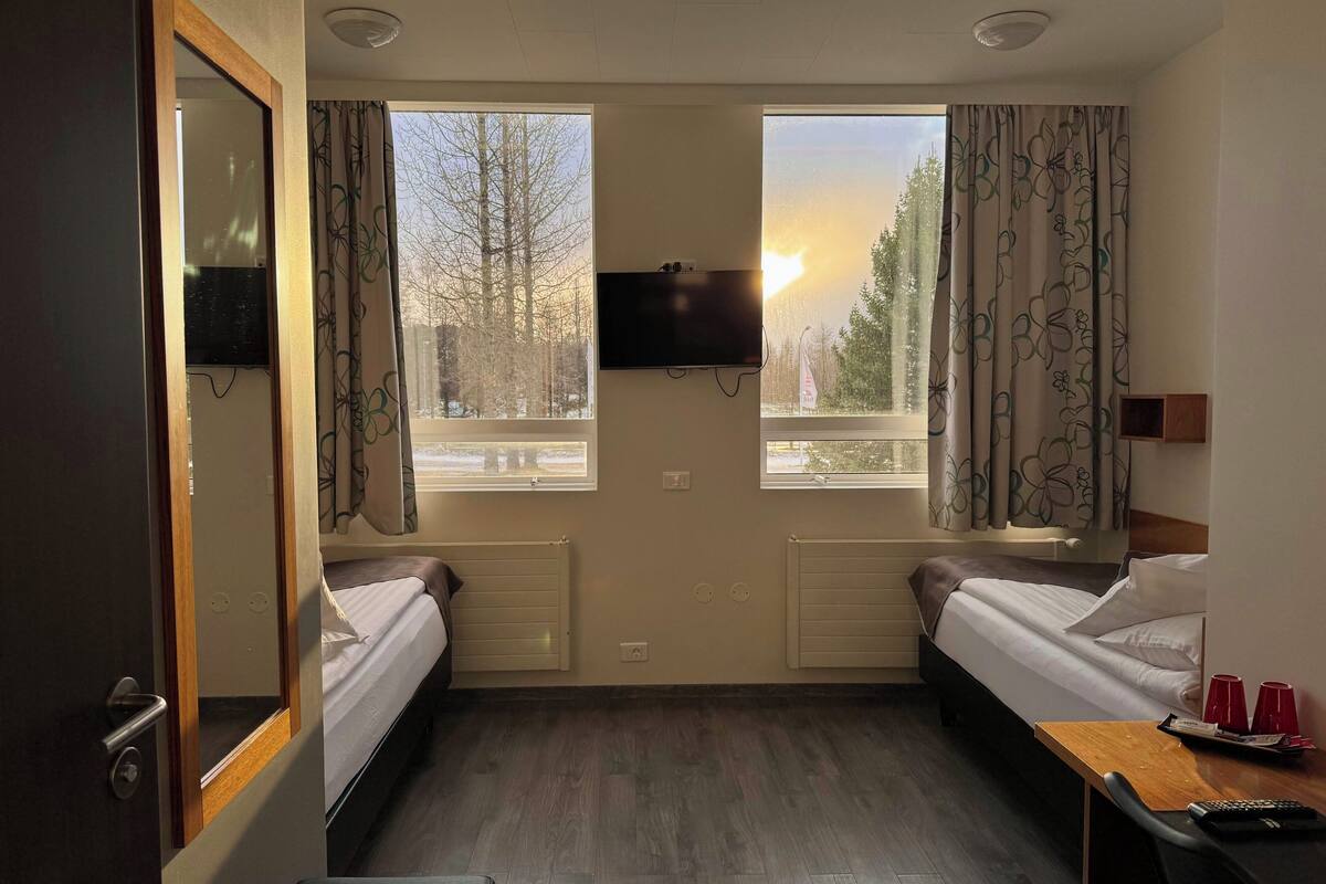 Ein Hotelzimmer mit zwei Betten, einem Fernseher, einem Fenster mit Blick auf Bäume und einem Holztisch mit Tassen und Tablett.