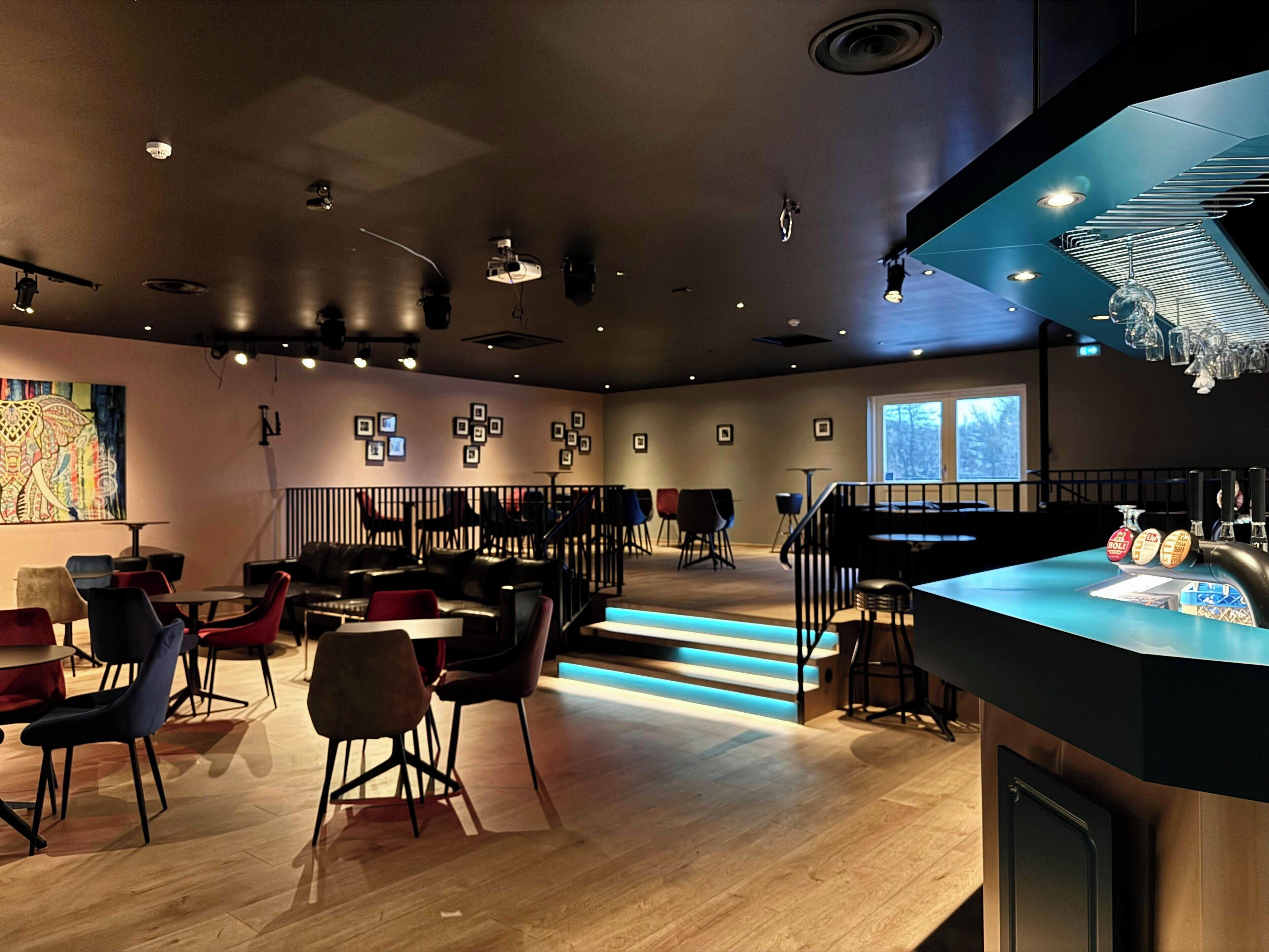 Un espace bar moderne doté d’un comptoir, de tabourets et d’un escalier menant à un espace salon avec des tables et des chaises.