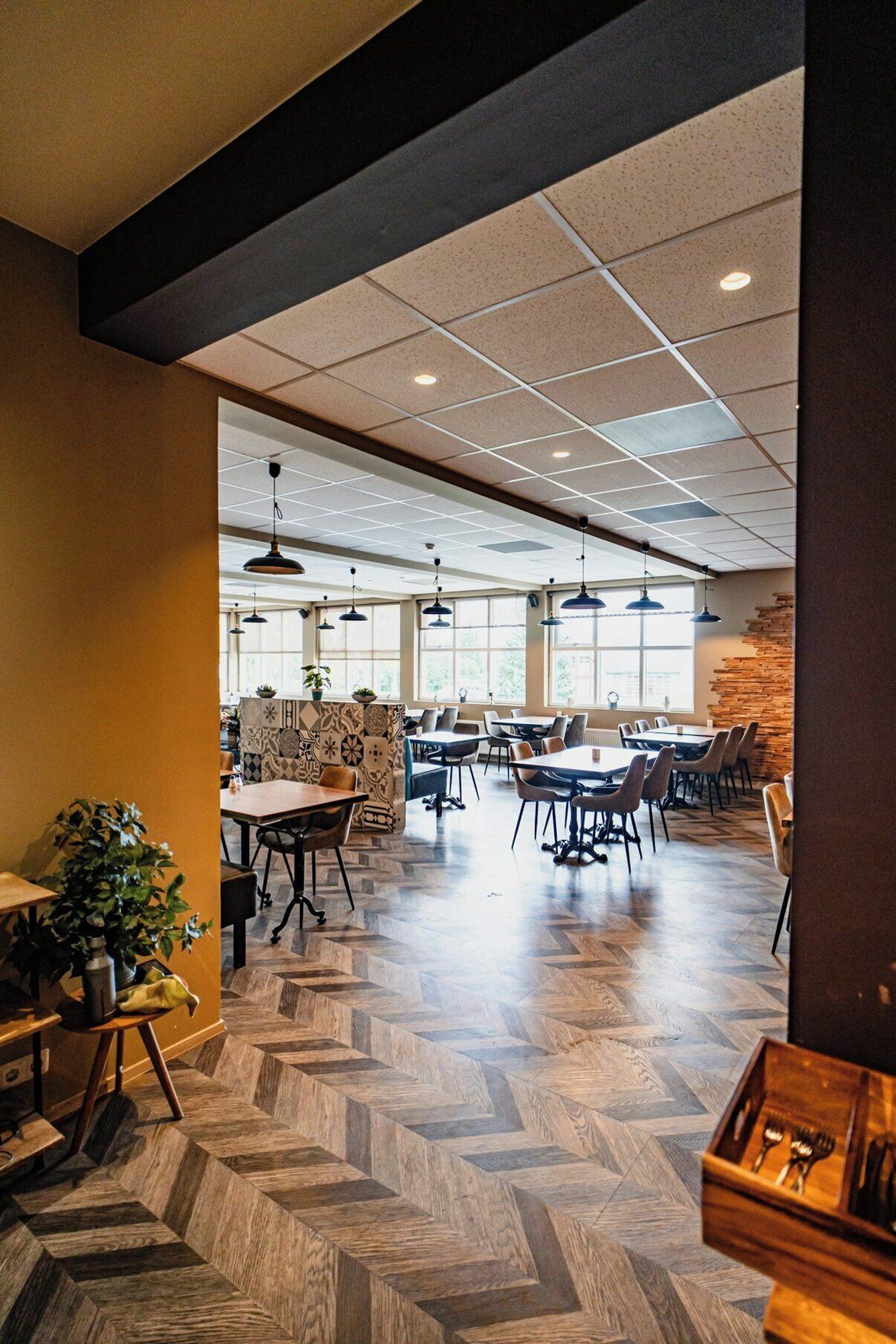 A spacious dining area with multiple tables and chairs, a herringbone-patterned floor, and a large window providing natural light.