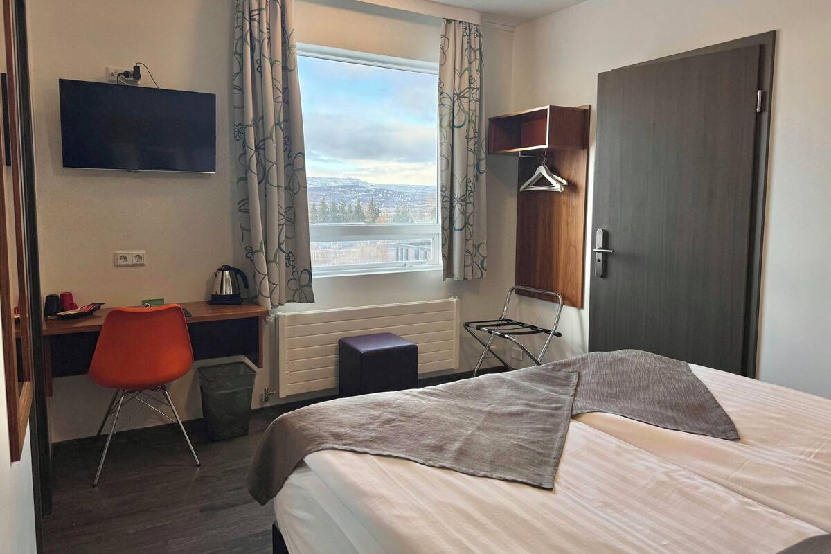 Ein Hotelzimmer mit Bett, Schreibtisch, Stuhl, Heizkörper, Fernseher und einem Fenster mit Vorhängen.