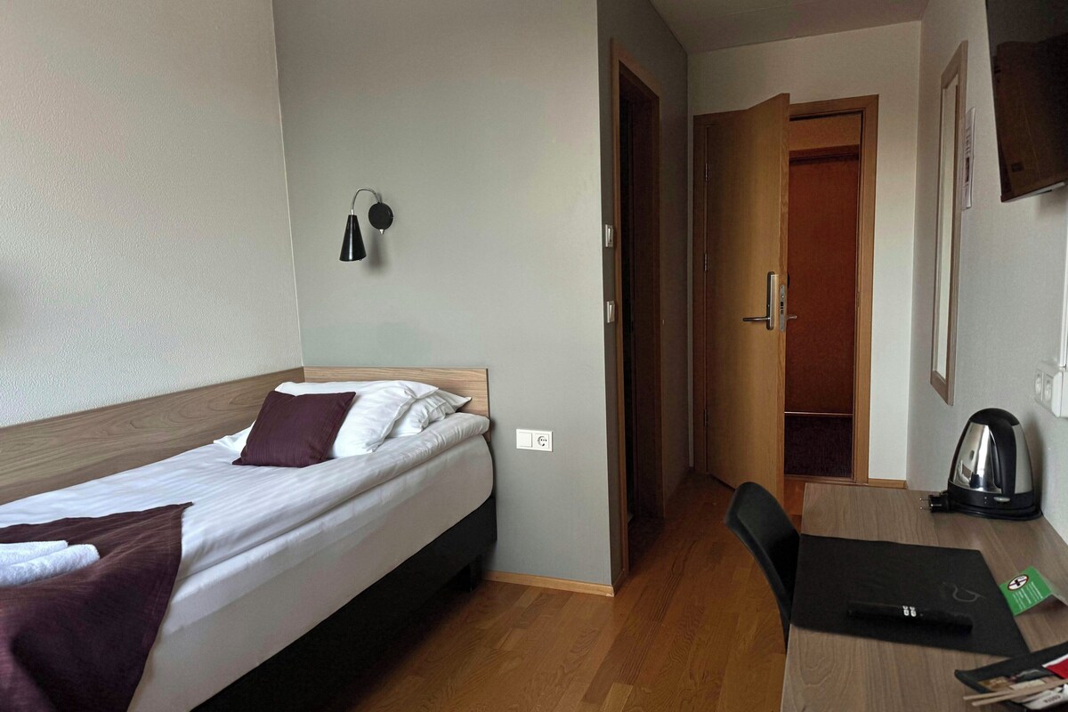 A hotel room with a bed, a desk, a chair, a coffee maker, and a door leading to another room.