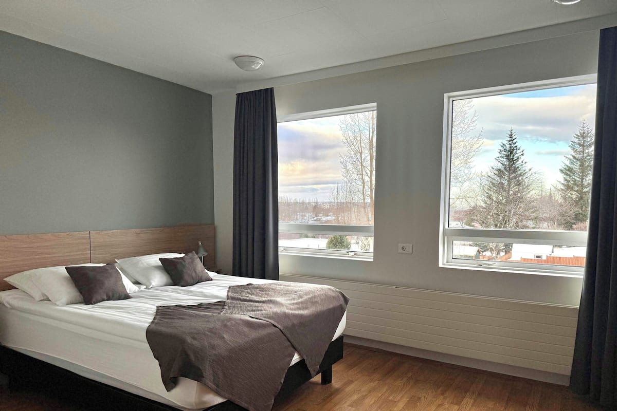 Ein modernes Hotelzimmer mit einem großen Bett, zwei Fenstern mit Vorhängen, einer Deckenleuchte und Blick auf Bäume und Gebäude.