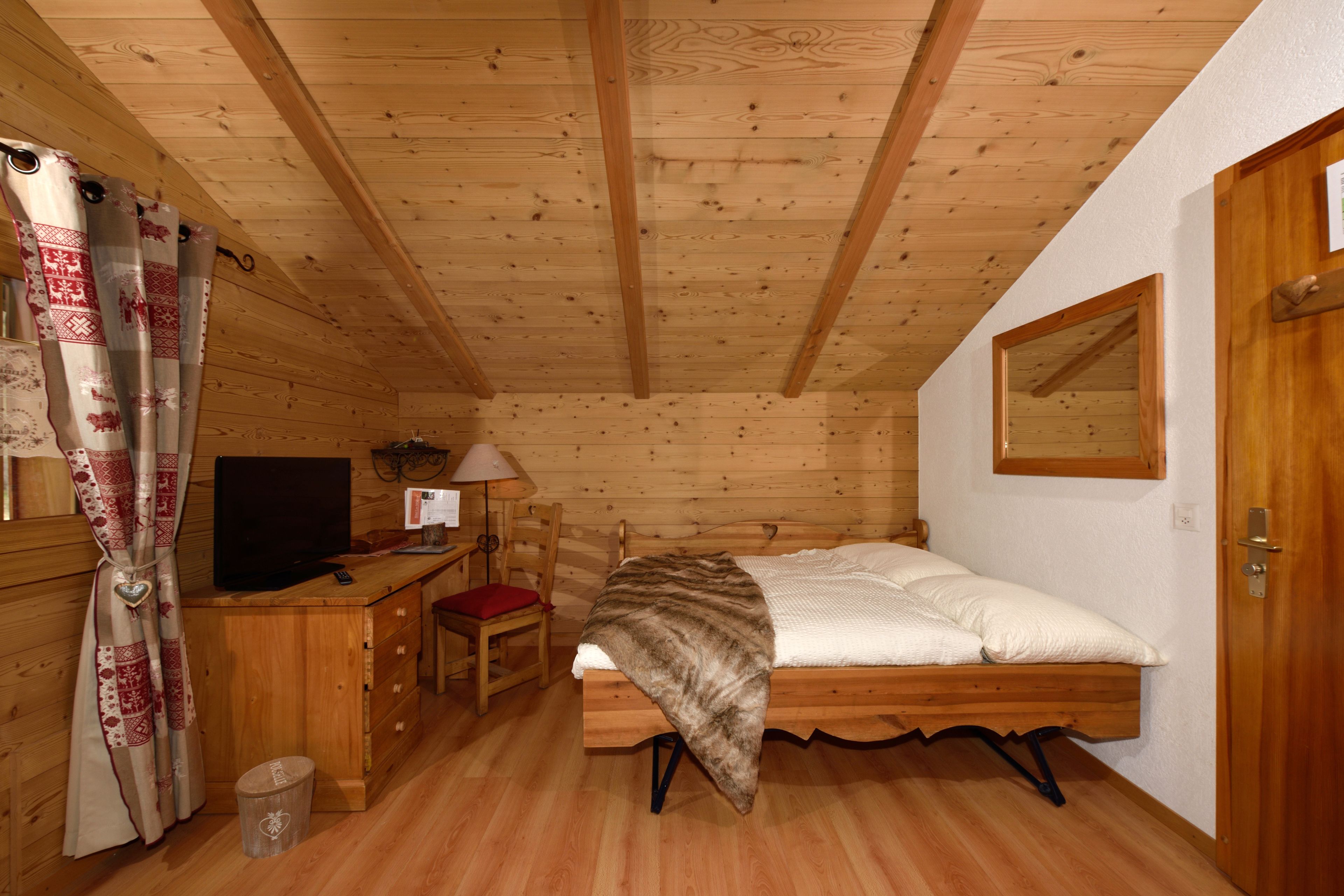 A cozy room with a wooden ceiling, a bed with a blanket, a desk with a computer, a chair, and a TV.