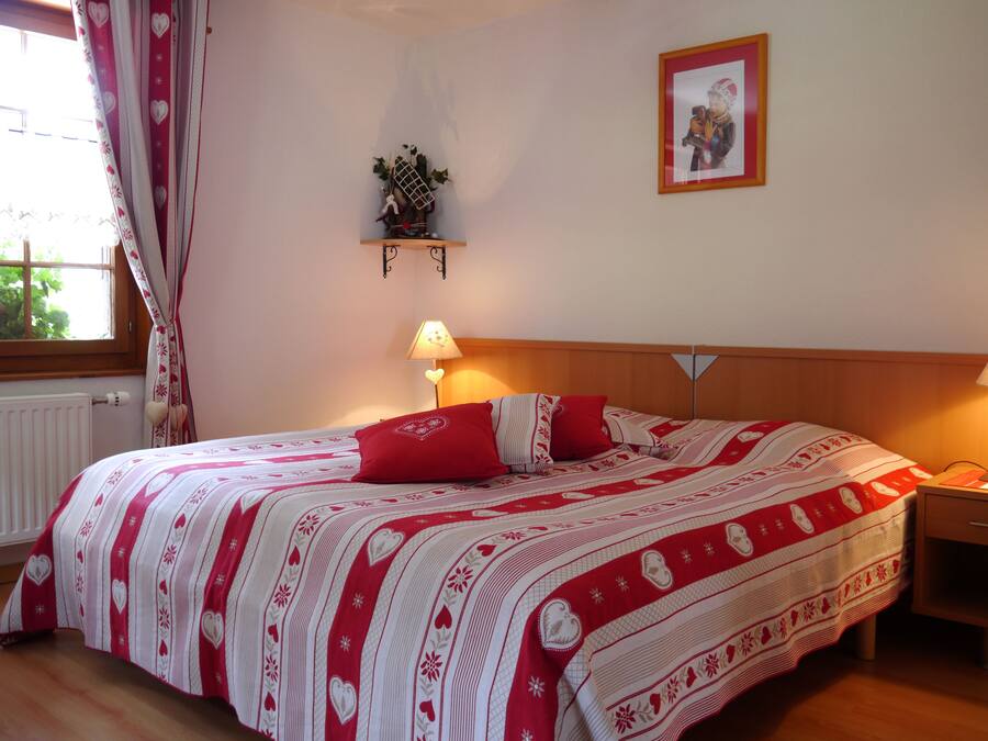 A bedroom with a large bed, two bedside lamps, a nightstand, a window with curtains, a wall-mounted shelf with a plant, and a framed picture on the wall.