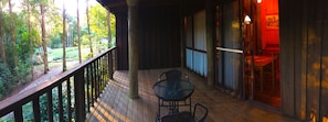 A wooden deck with a glass table and chairs, overlooking a garden with trees and a small building. - Ecostudio Fellini (Mudgeeraba)