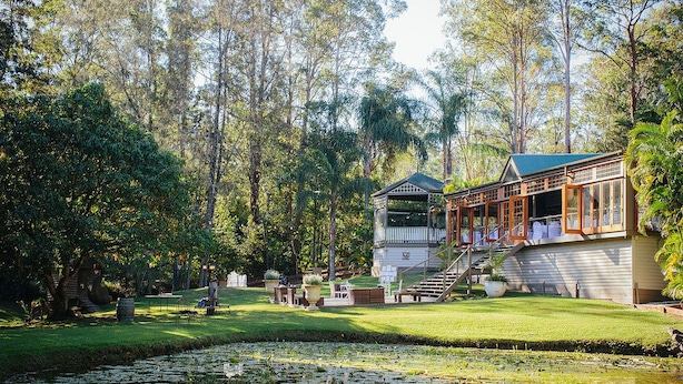 Um edifício com varanda coberta e escadaria para um deck, cercado por um lago e árvores densas.