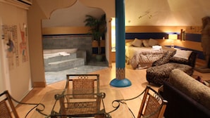 Quarto de hotel com uma cama, dois sofás, uma mesa de centro e uma cadeira. Há uma escada que leva a uma varanda com uma planta.